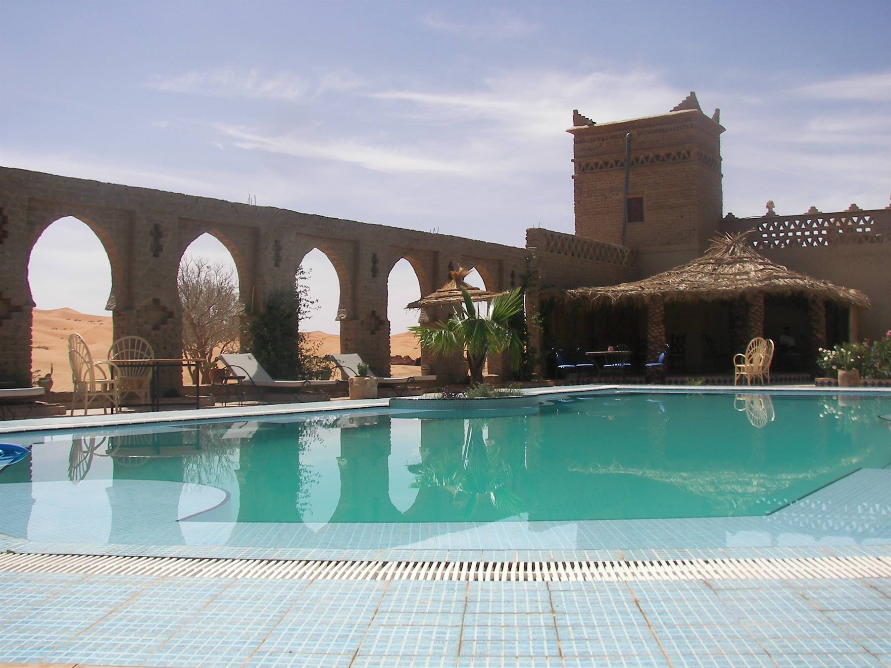 Blue pool in front of adobe kasbah with arches, palms and dunes