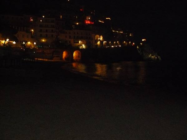 Dark beach at night, lit houses on the cliff reflect on the sea