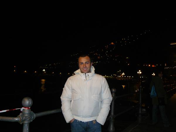 Man in white jacket poses at night beside an iron railing on the waterfront