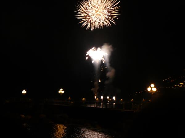 Golden fireworks explode at night over the lit seaside promenade, reflected on the sea