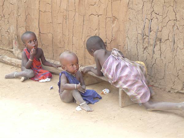 Maasai children sitting on a wooden bench in front of the mud wall