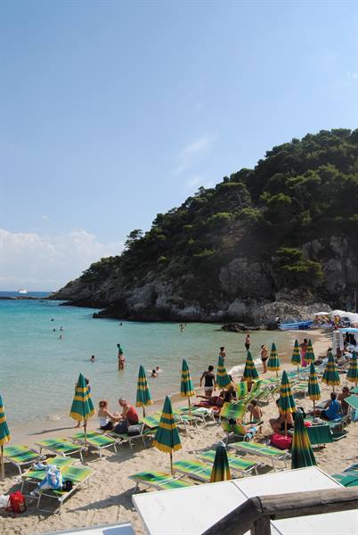 Beach with green and yellow umbrellas, swimmers in turquoise water below wooded hill