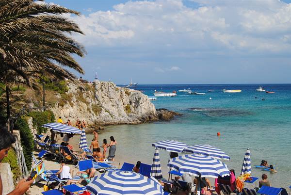 Blue striped umbrellas on the beach, rocky cliff with palms, boats on turquoise sea
