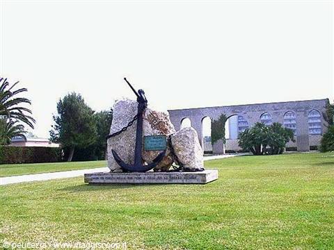 Memorial with black anchor and chain on rocks, green lawn and aqueduct arches behind