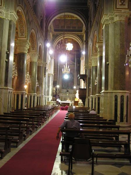 Church nave with marble columns, wooden pews, red carpet and lit chandeliers