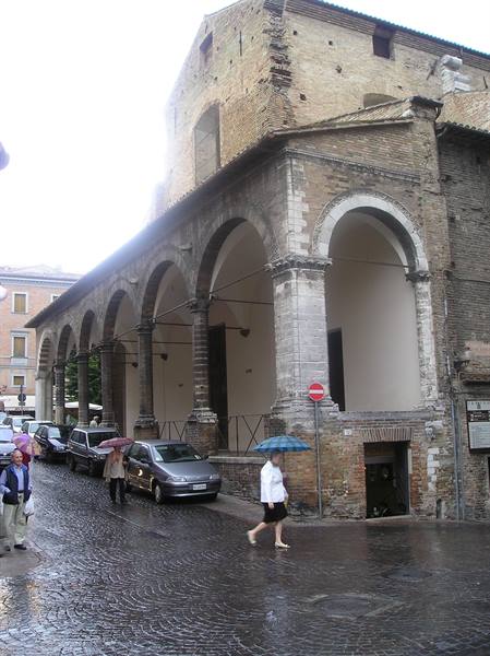 People with umbrellas walk past medieval brick arcades on a rainy cobblestone street