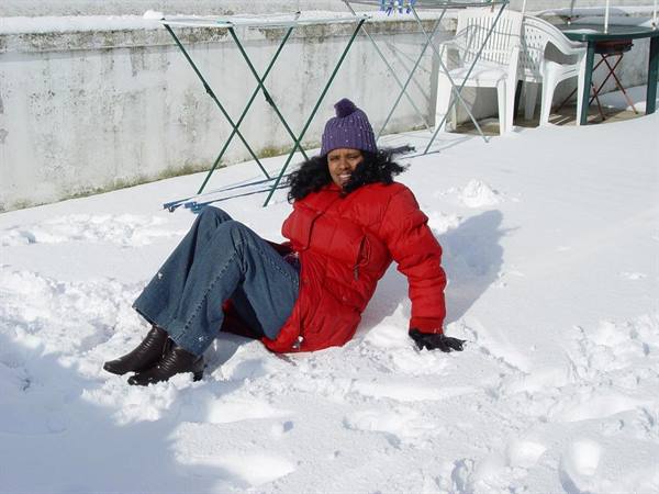 Woman in red coat and purple hat sitting smiling on snow, snowy terrace behind