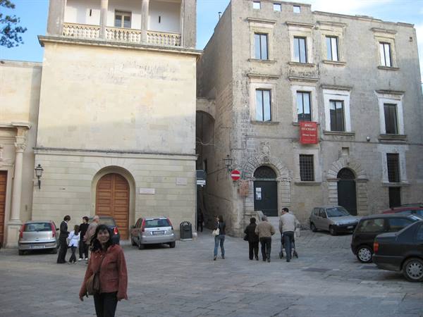 Woman in red jacket walks across square with stone buildings and cars