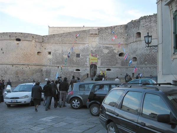 People and cars in front of a stone castle walls with flags