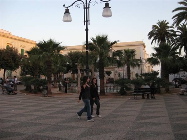 Two girls walk across a paved square with palm trees and pale buildings