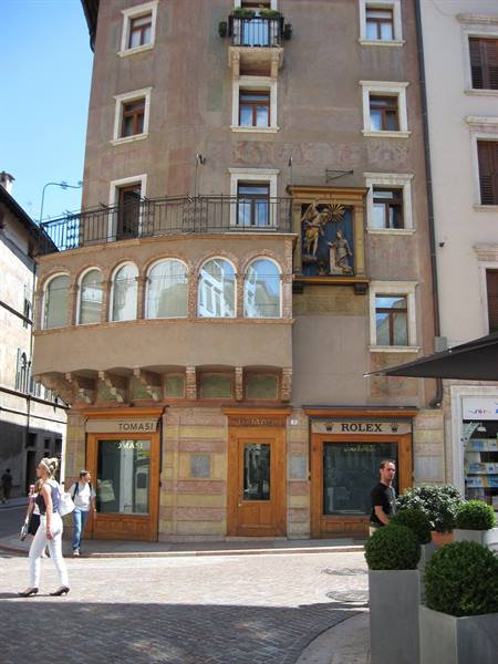 Stuccoed corner building with painted shrine above shops, pedestrians in sunlight on cobblestones