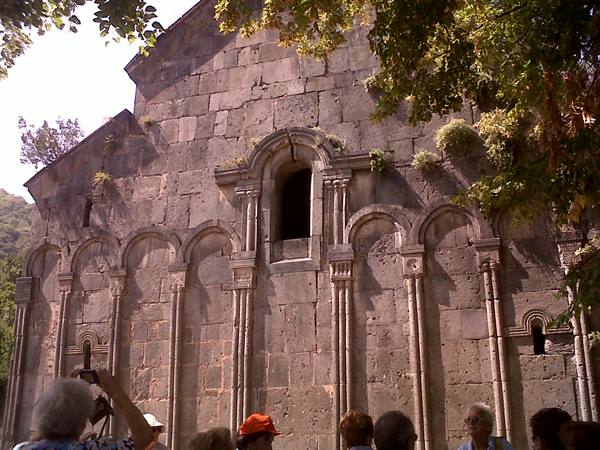 Tourists photograph the grey stone facade with blind arches among trees