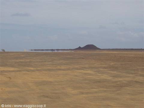 Flat sandy desert plain with dark volcanic hill on the horizon, overcast sky