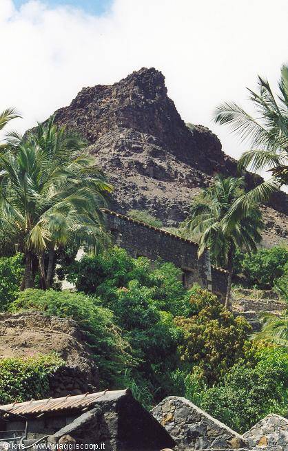 Volcanic crag above stone houses and palms, old wall among lush greenery
