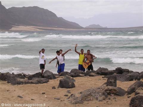 Boys with guitar on rocky beach, waves crashing, mountains in background