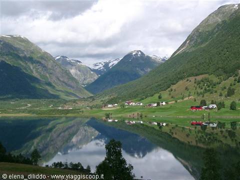 Red farmhouses and green meadows reflect in the lake beneath snowy mountains