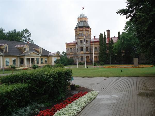 Sigulda New Castle among trees, red flowerbeds and wet paved path under grey sky