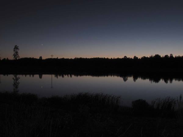 Dark lake reflecting trees and pole, orange sky with star at dawn
