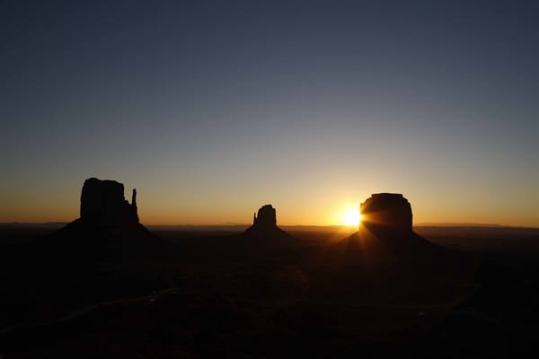 Sunrise behind Monument Valley buttes, dark silhouettes rising over the flat desert