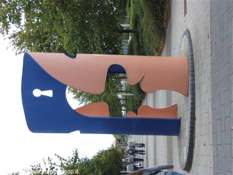 Blue and pink metal sculpture with stylized faces along a tree-lined sidewalk
