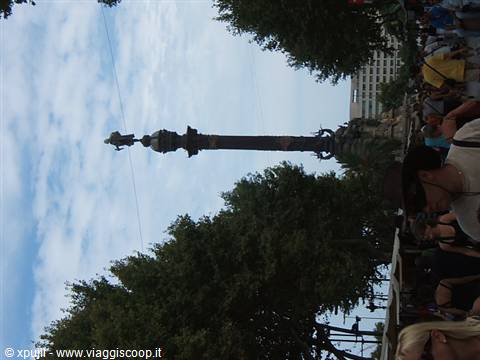 Column with Columbus statue among trees, tourists nearby, cloudy sky
