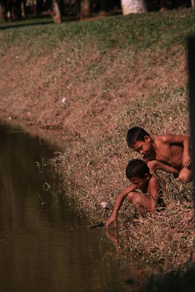Two shirtless boys touch the pond water from the grassy bank