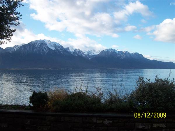 Lake Geneva with snow-capped Alps among clouds, plants on stone wall
