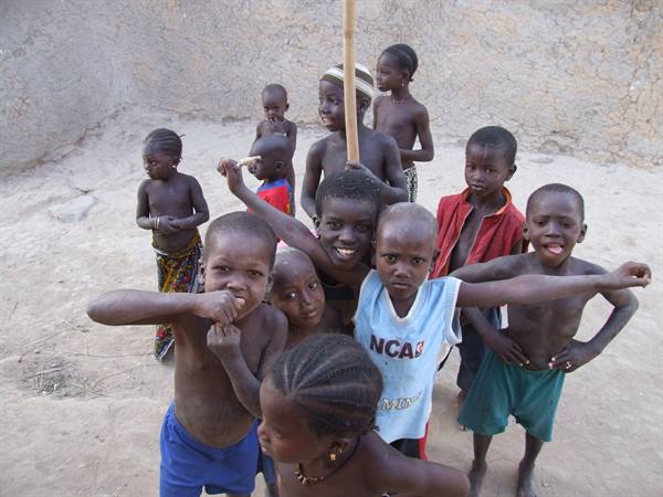 Smiling children group in a dirt courtyard, one holding a wooden stick