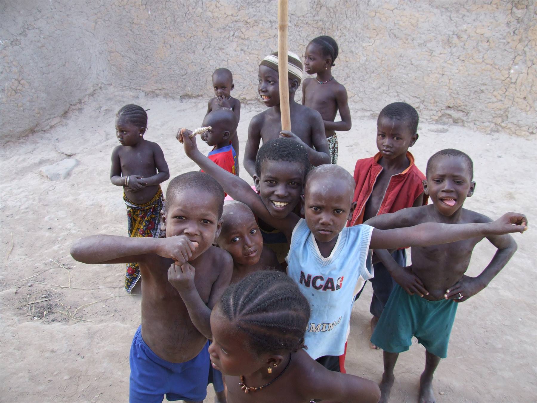 Smiling children group in a dirt courtyard, one holding a wooden stick