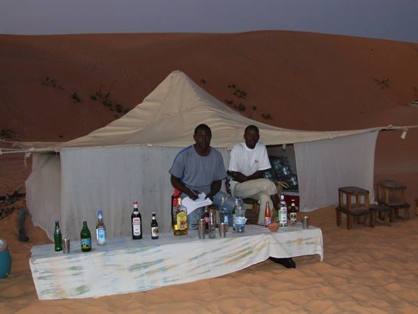 Two men behind a tent-camp bar amid the sand dunes