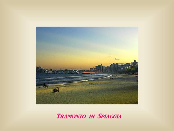 Two people sitting on sand facing ocean and buildings lit by low sun