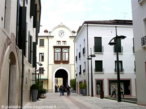 Pedestrians on a cobbled square between pale buildings with clock and arched gateway