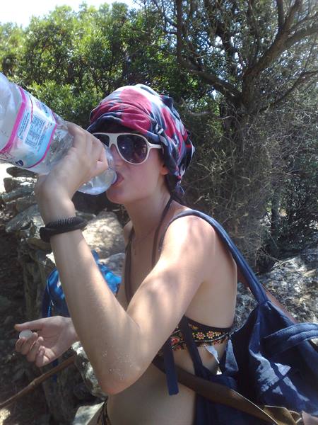 Girl in headscarf and sunglasses drinking water among Mediterranean bushes and stone walls