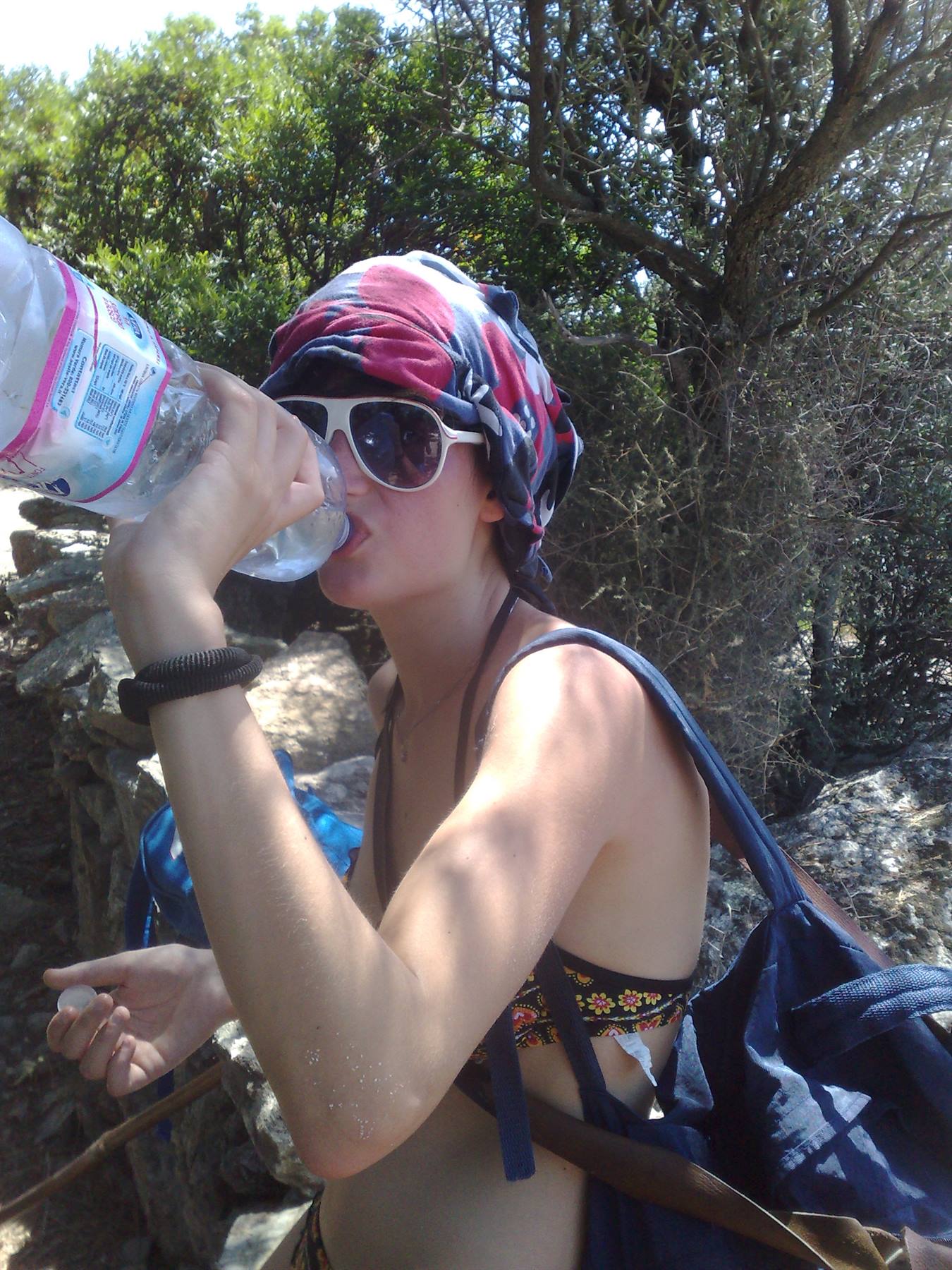 Girl in headscarf and sunglasses drinking water among Mediterranean bushes and stone walls