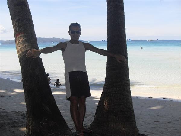Man in tank top leaning between two palm trunks on white beach with sailboats