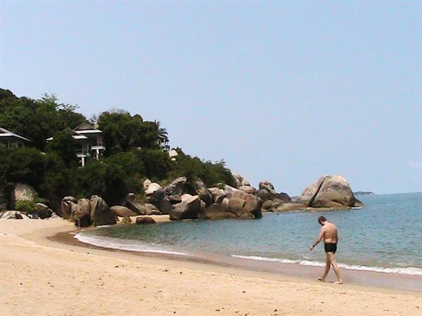 Man in swim trunks walking on sand beside large boulders and hillside bungalows
