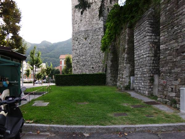 Medieval stone walls with tower, green lawn below, parked scooter, mountains in background