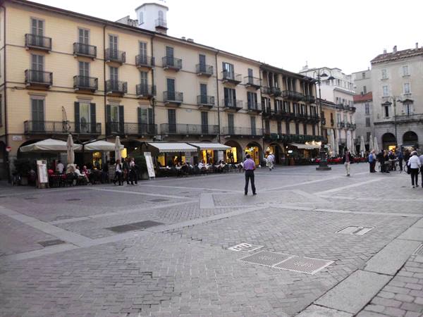 Square with cafés under arcades, people stroll across cobblestones at dusk