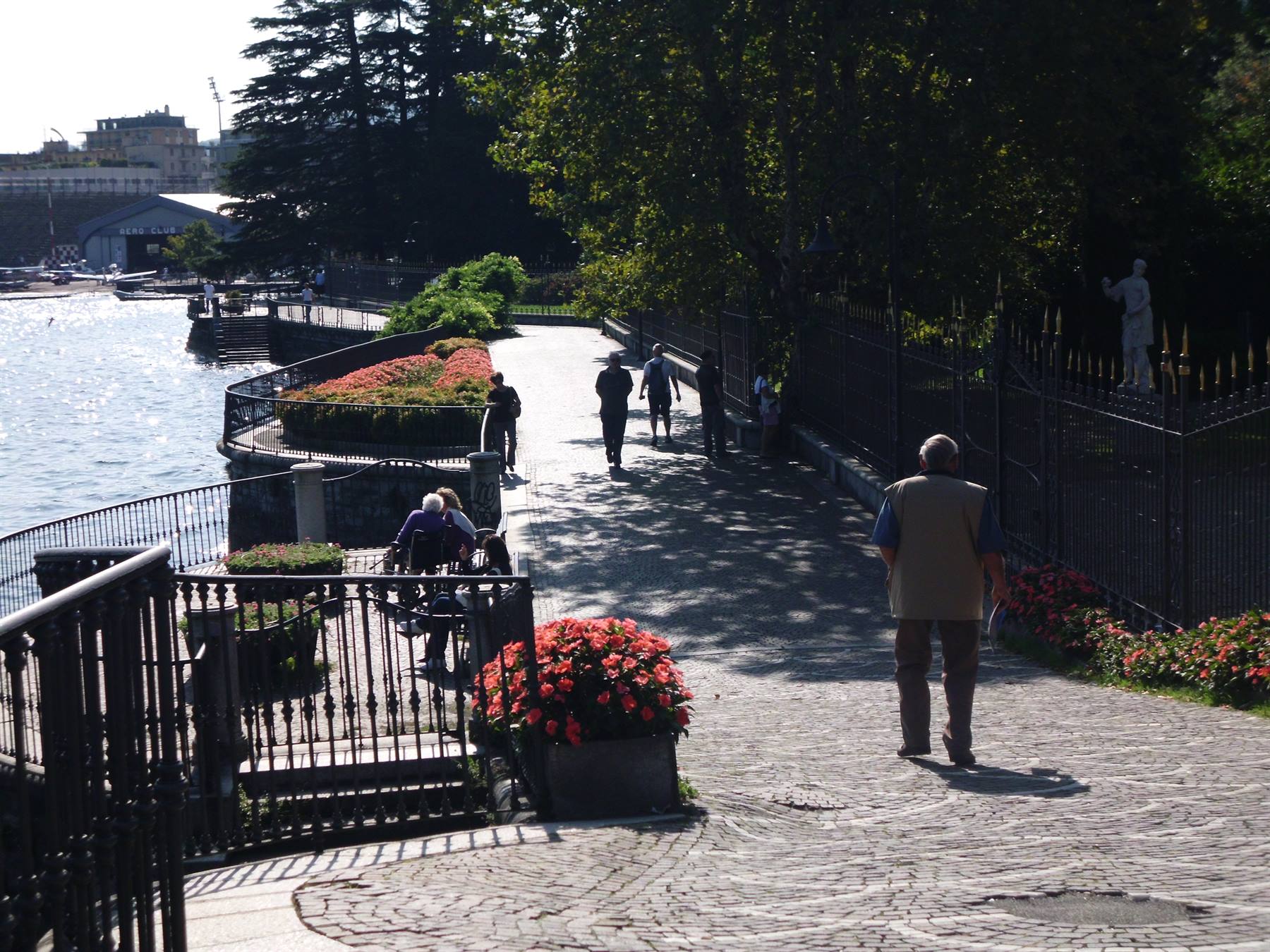 Stone lakeside promenade, red flowers, iron railings, people walking in sunlight