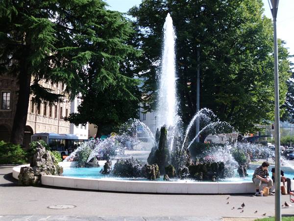 Rocky fountain with water jets in a tree-lined square, people sitting on the edge