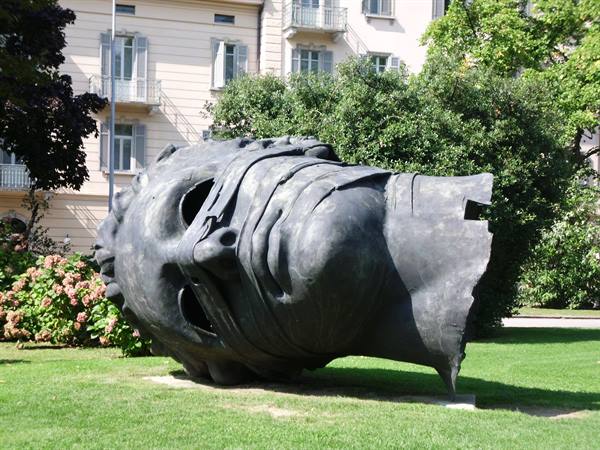 Large fractured bronze head by Mitoraj lying on the lawn, trees and buildings behind