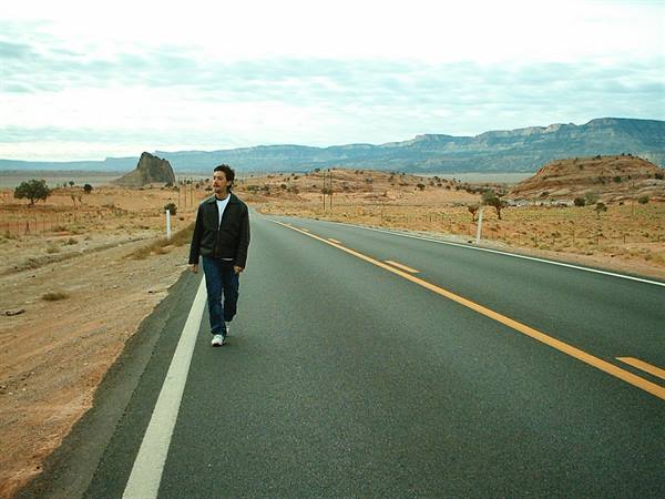Man walking on asphalt desert highway, red rocks and mesas under clouds