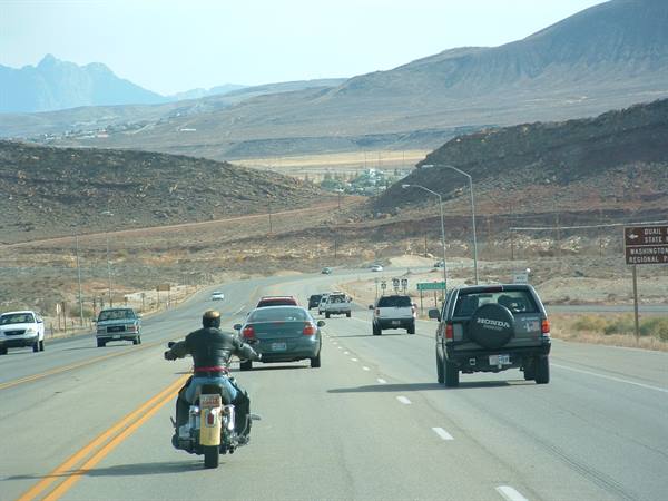 Leather-jacketed motorcyclist rides among cars on desert highway past arid hills