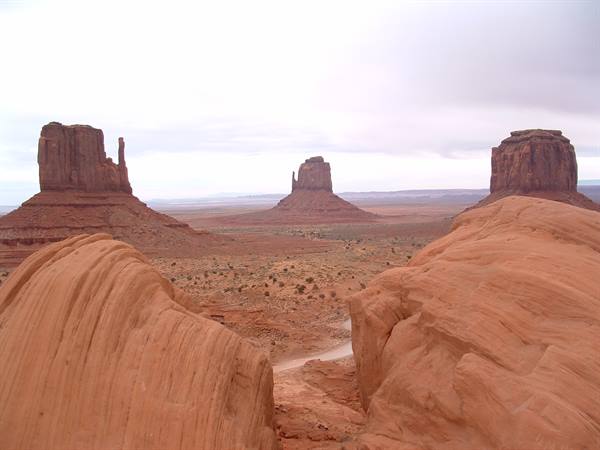 Monument Valley: red sandstone buttes under cloudy sky, sandy desert floor with shrubs