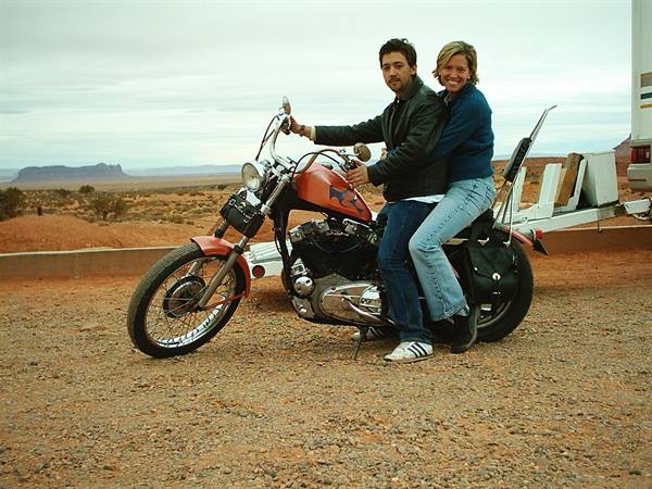 Couple on orange motorcycle over red gravel, Monument Valley under cloudy sky