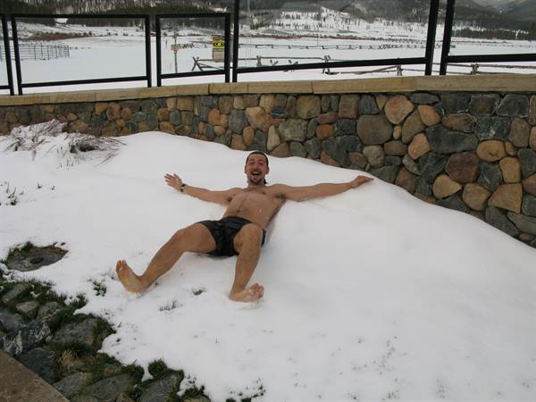 Man in swim shorts sprawls on snow with open arms by a stone wall