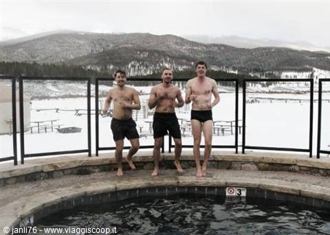 Three men in swimwear at hot tub edge, snow and mountains behind