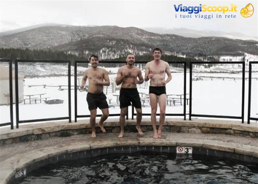 Three men in swimwear at hot tub edge, snow and mountains behind