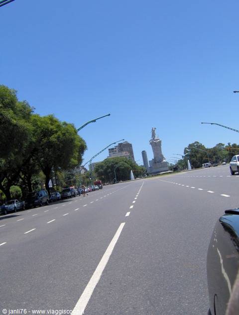 Wide avenue with monument, street lamps and green trees under blue sky