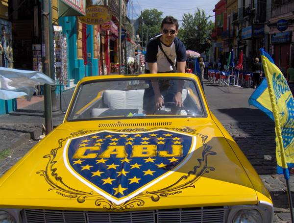 Man on yellow car with Boca Juniors crest, cobbled street of El Caminito
